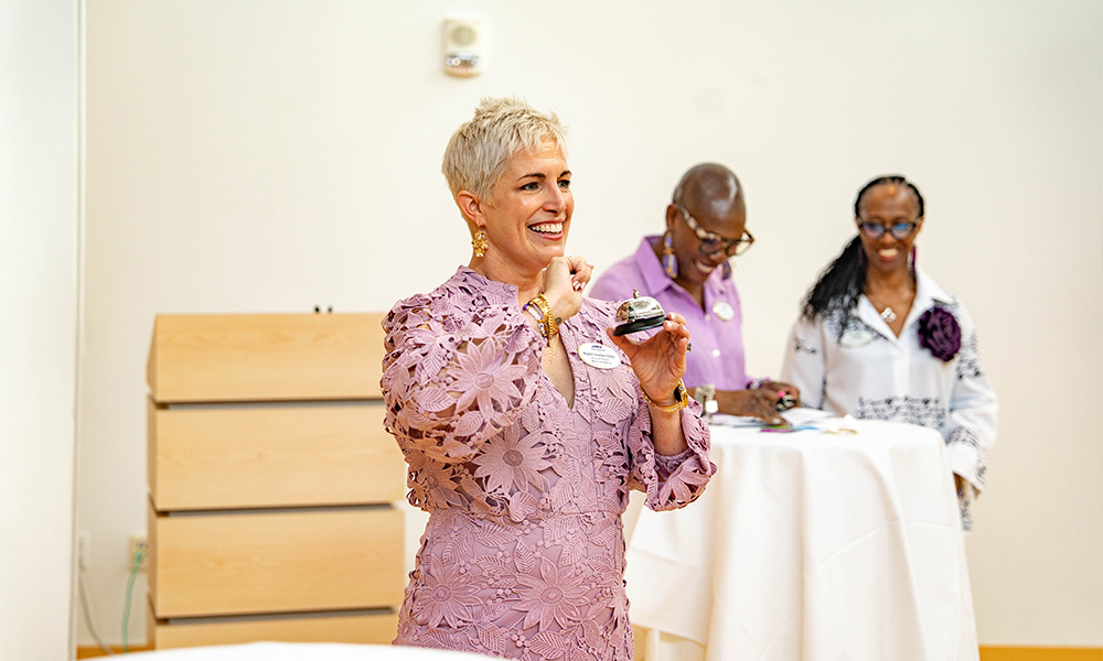 A smiling woman in a lavender dress holds a small object while speaking, with two other women in the background engaged in conversation.