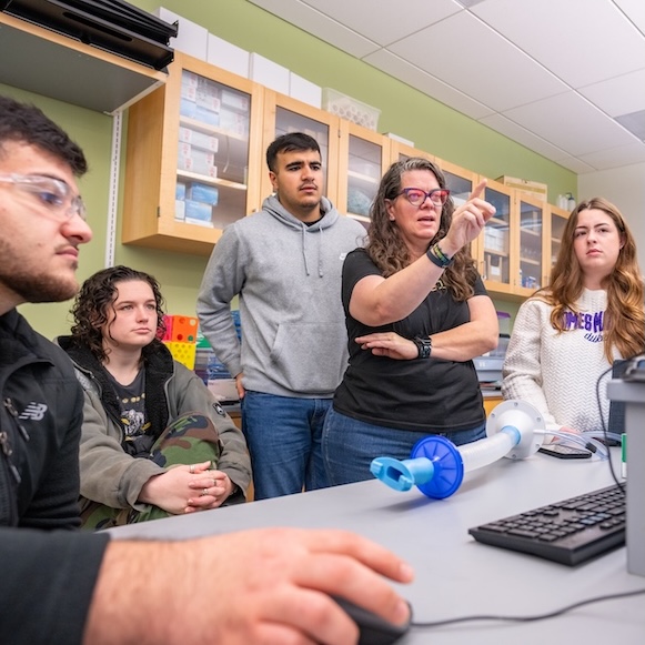 Instructor points at a computer screen while students watch in a science lab classroom.