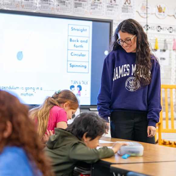 Teacher in a classroom smiles while students work at a table, with a lesson display on motion in the background.