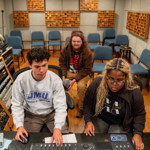 Two people operate an audio mixing console in a recording studio while a third person watches from behind.
