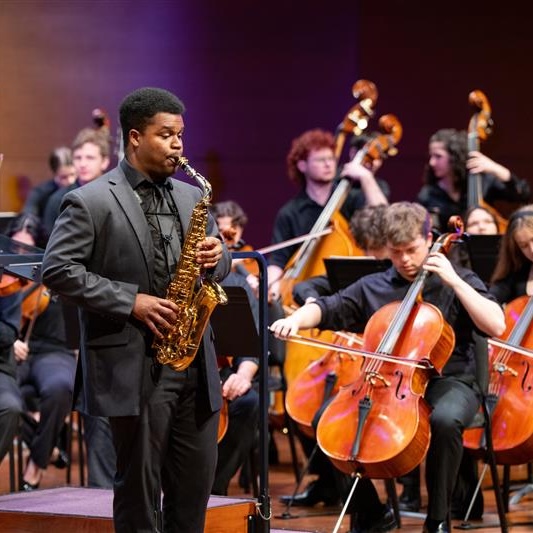 A saxophonist performs solo in front of an orchestra during a concert.