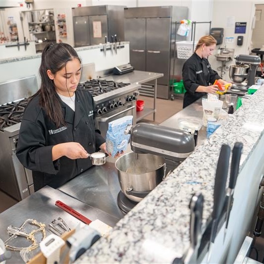 Two culinary students working in a professional kitchen, one measuring ingredients while the other prepares dough.