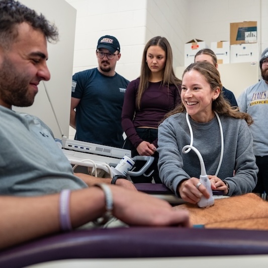 A group of students observes as a woman conducts a hands-on demonstration with a patient in a medical training setting.