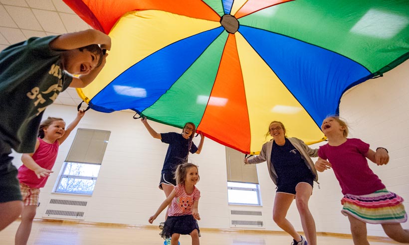 Kinesiology students and children practice motor skills under a colorful parachute