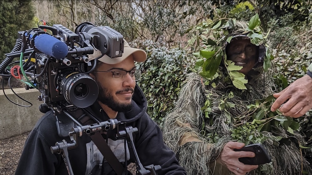 A man operating a camera sits in the foreground while a person in camouflage made of leaves and branches blends into the background.