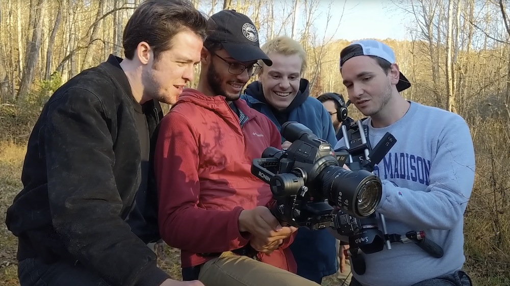 Four young men gathered around a camera, reviewing footage together in an outdoor setting.