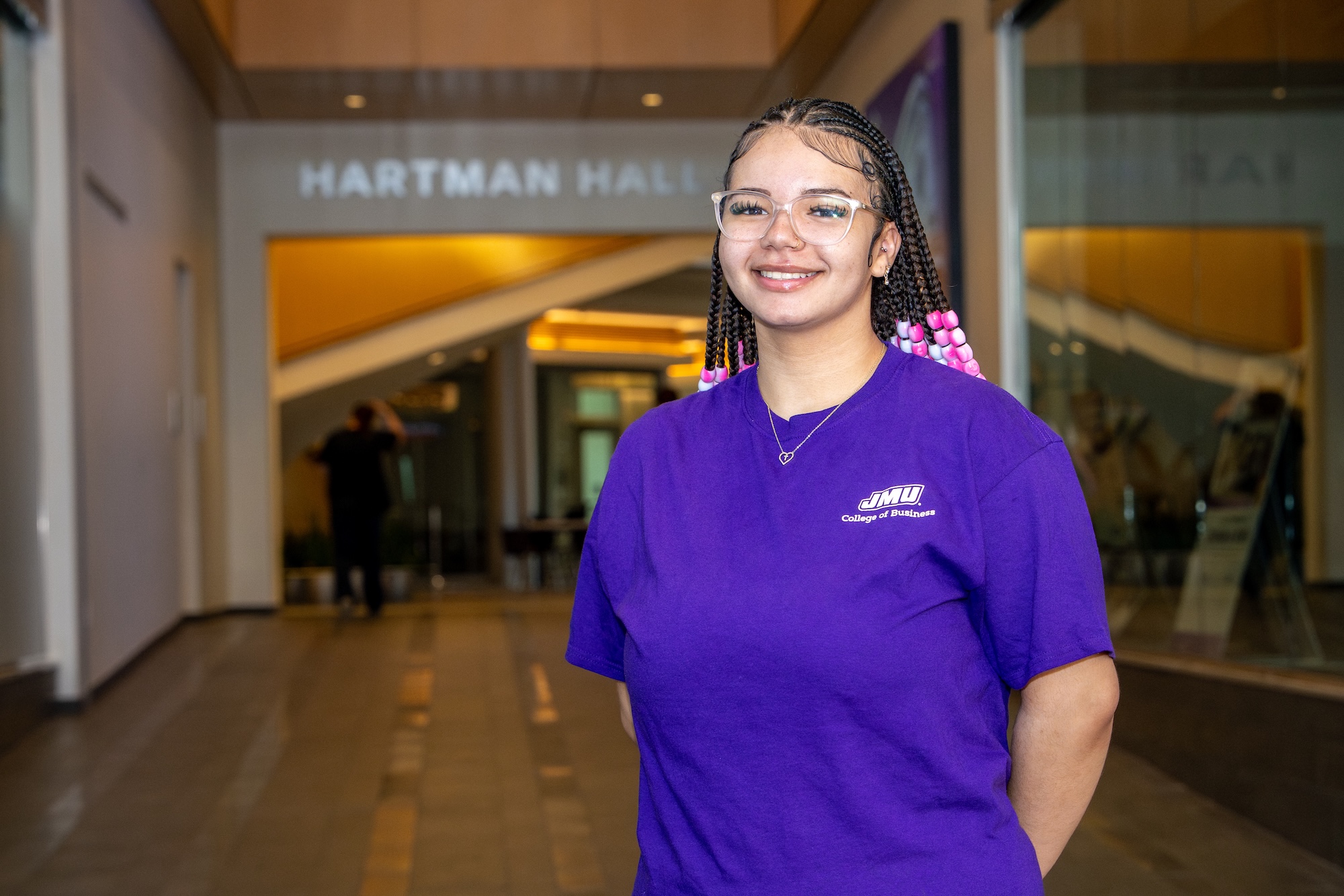 Bryanna Hill smiling in a purple JMU College of Business T-shirt leaning on a railing in an indoor campus building.