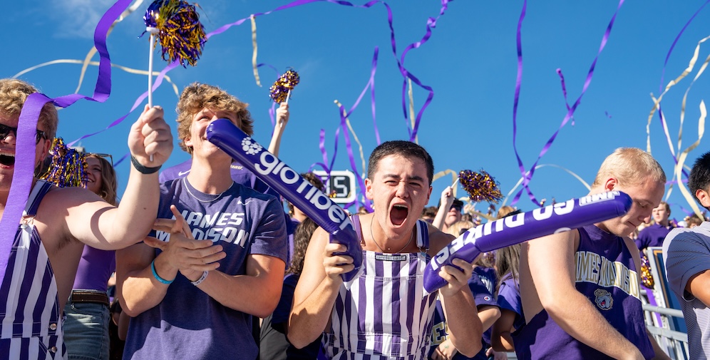 Cheering fans in purple attire celebrate at a sporting event, waving pompoms and noise makers in a spirited display of team pride.