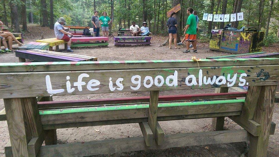 students at a summer camp. In the foreground, a bench painted with the slogan "Life is good. Always."