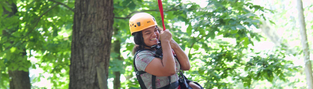 A Camp Easterseals camper wears a harnessa and helmet and smiles while going down a zipline