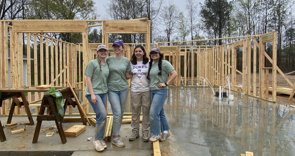 Three Alternative Spring Breakers pose in front of a construction site, with the wooden frame of a house in the background.