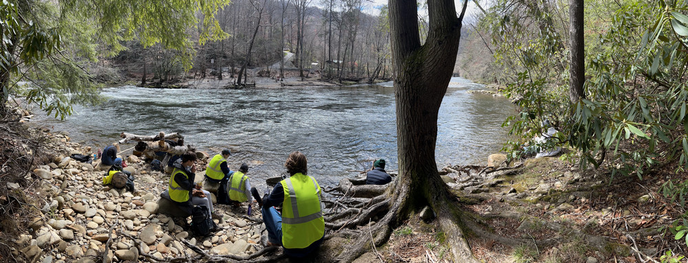 The 2023 Great Smoky Mountains group rests in front of a river while wearing neon safety vests mountain vista in the background