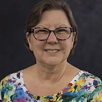 Smiling woman wearing tortoiseshell glasses and a colorful floral top against a dark studio background.