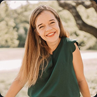 Smiling woman with long hair in a dark green sleeveless top outdoors, with sunlight and trees in the background.