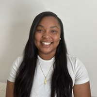 Smiling person with long dark hair wearing a white T-shirt, photographed against a light gray background.