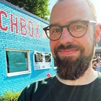 Close-up selfie of a bearded man wearing glasses, with a bright blue wall and partial red lettering in the background.