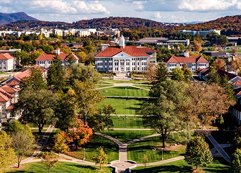 Aerial view JMU University campus with a central lawn, pathways, red-roofed buildings, and autumn trees, with hills in the background.