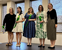 Five women stand side by side indoors, smiling for a group photo, with two holding bouquets of flowers.