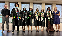 Group of graduates in black caps and gowns with green-and-gold stoles pose indoors with two faculty members at either end.