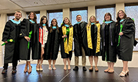 Group of graduates in caps and gowns standing indoors and smiling, with several holding green flowers.