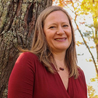 Cheri Greenfield, smiling, in a maroon top leaning against a tree trunk outdoors with autumn leaves in the background.