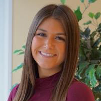 Madison Moore,  smiling young woman with long brown hair, wearing a burgundy top, stands in front of greenery.