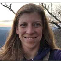 Smiling person with long hair outdoors at sunset, with mountains and bare tree branches in the background.