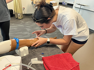 A JMU student practicing a medical procedure on a training arm model in a classroom setting.