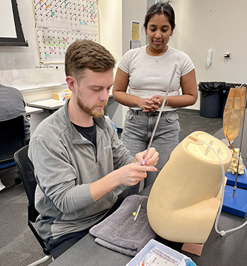 A man sitting at a table practicing a lumbar puncture on a prosthetic device while a woman stands nearby observing.