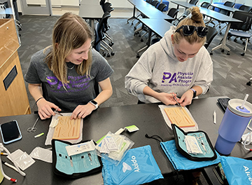 Two JMU students engaged in a hands-on suturing activity at a table, with various supplies around them.