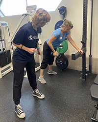 Two women engaged in strength training exercises in a gym, one lifting a dumbbell while the other prepares equipment.