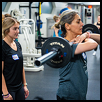 EmpowerHER - Two women participating in a weightlifting session at a gym, with one woman lifting a barbell while the other observes.