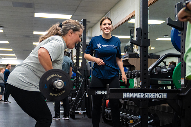 EmpowerHER - Two women are engaged in a strength training session at a gym, with one lifting weights and the other encouraging her.