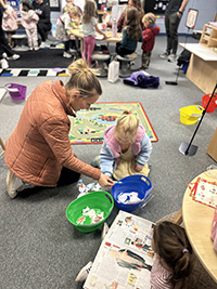 A parent helps a child with an art project in a colorful classroom filled with engaged children participating in various activities.