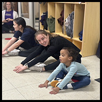 Healthy Kids program - Two older children and a young girl are stretching on the floor in a classroom setting, engaging in a playful exercise activity.