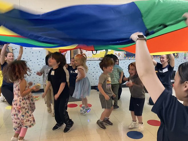 Healthy Kids - Children joyfully playing under a colorful parachute in a classroom setting, surrounded by adults who are facilitating the activity.