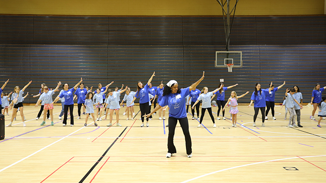 Movin and Grovin Day - A group of people in blue shirts dance together in a gymnasium, with one leader guiding the activity in front.