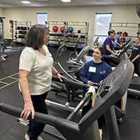 A fitness instructor guiding a participant on a treadmill in a gym setting, with various exercise equipment and other attendees in the background.