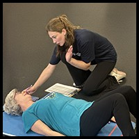 Older Women's Wellness event - A trainer demonstrates a health assessment with an older woman lying on an exercise mat.