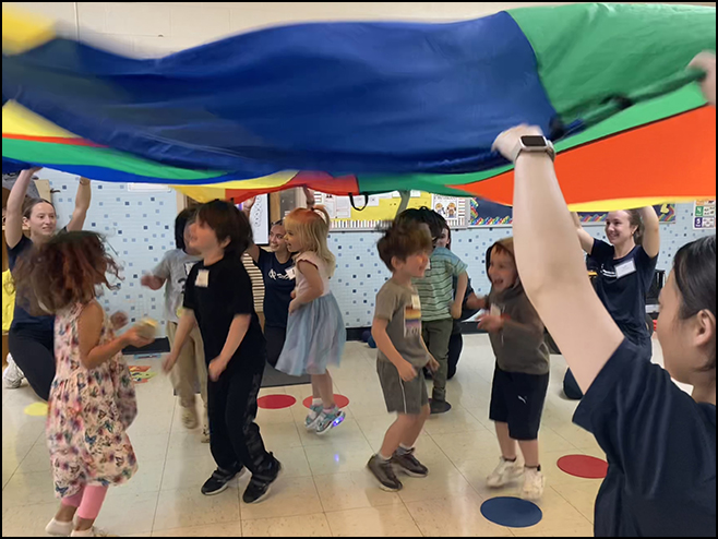 Healthy Kids - Children joyfully playing under a colorful parachute in a classroom setting, surrounded by adults who are facilitating the activity.