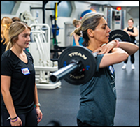 EmpowerHER - Two women participating in a weightlifting session at a gym, with one woman lifting a barbell while the other observes.