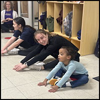 Healthy Kids program - Two older children and a young girl are stretching on the floor in a classroom setting, engaging in a playful exercise activity.