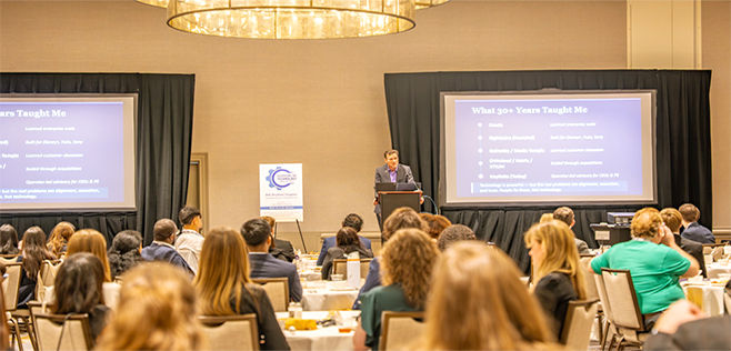 Speaker presenting at the AIS conference to a seated audience, with a slide deck displayed on two large screens.