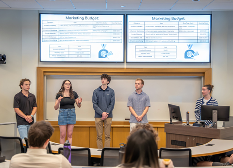 Students making a presentation on stage in a classroom setting, with a projected image of a table showing categories and figures.