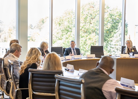 A group of people are seated around a large, curved conference table in a modern meeting room. The table is equipped with nameplates, papers, and water bottles. Several individuals are visible in the foreground with their backs to the camera, while three people are clearly seen facing the camera across the table. Behind them, tall floor-to-ceiling windows reveal a bright outdoor scene with trees in autumn colors. The room is well-lit with natural light, and the chairs are sleek, high-backed office chairs arranged around the table.