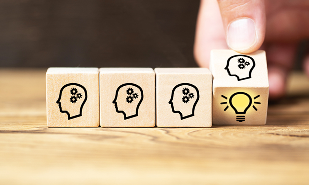The image shows four wooden blocks placed in a row on a wooden surface. Each block has a black outline of a human head in profile with two gear icons inside, symbolizing thinking or ideas. The first three blocks are identical, showing just the head with gears. The fourth block, which is being held by a hand, also has the head with gears but includes a bright yellow light bulb icon below it, symbolizing innovation or a new idea. The background is blurred, keeping focus on the blocks.