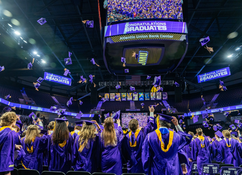 JMU Graduates in purple gowns toss their caps in an indoor arena, with a large scoreboard displaying “Congratulations Graduates.”