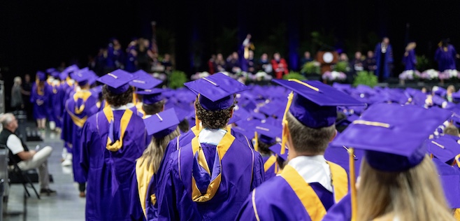 College of Business graduates in purple caps and gowns line up at a commencement ceremony, with a crowd and stage in the background.