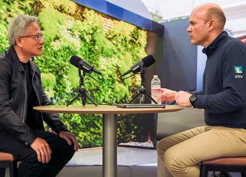 Jensen Huang and Eric Veiel sit across from each other at a small round table with microphones, recording a podcast or interview in front of a green plant wall backdrop.