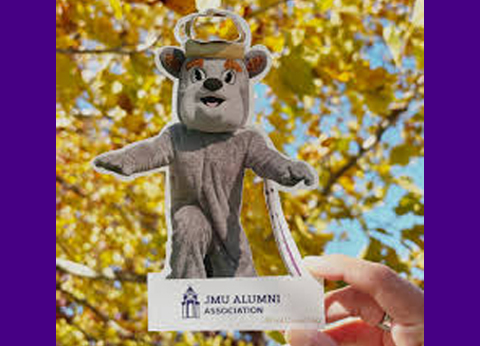 Cutout of the Duke Dog mascot held up against yellow autumn leaves, with “JMU Alumni Association” printed on the base.